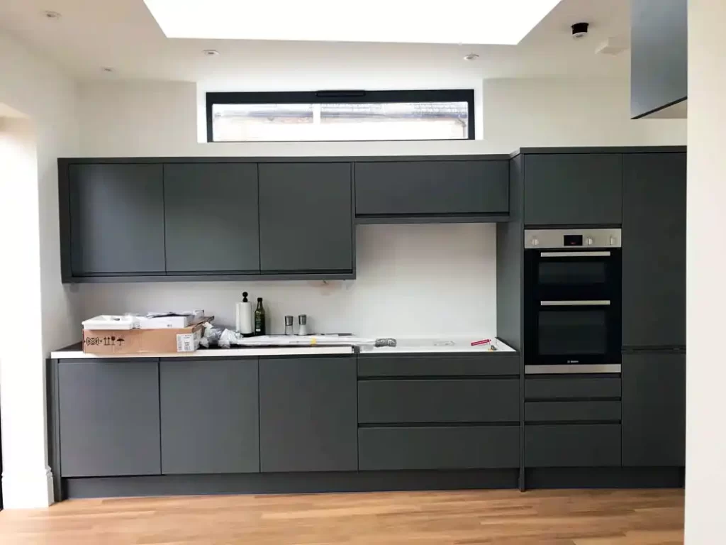 Modern fitted kitchen with matte dark-grey handleless cabinets, built-in double oven and a long worktop beneath a wide rooflight and high-level window.